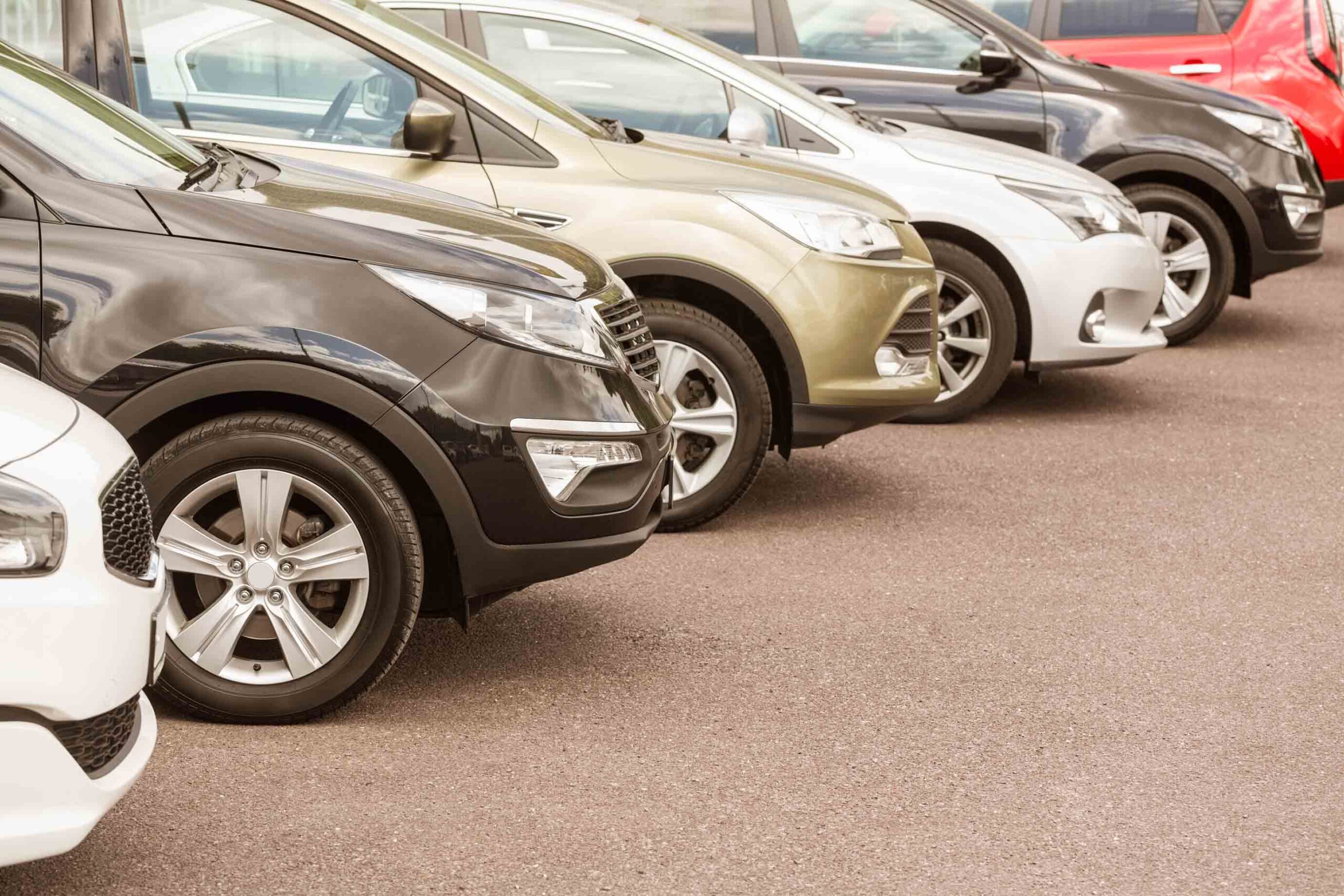 A diagonal row of parked cars lined up on an asphalt surface, shown from a low angle focusing on their front ends and alloy wheels